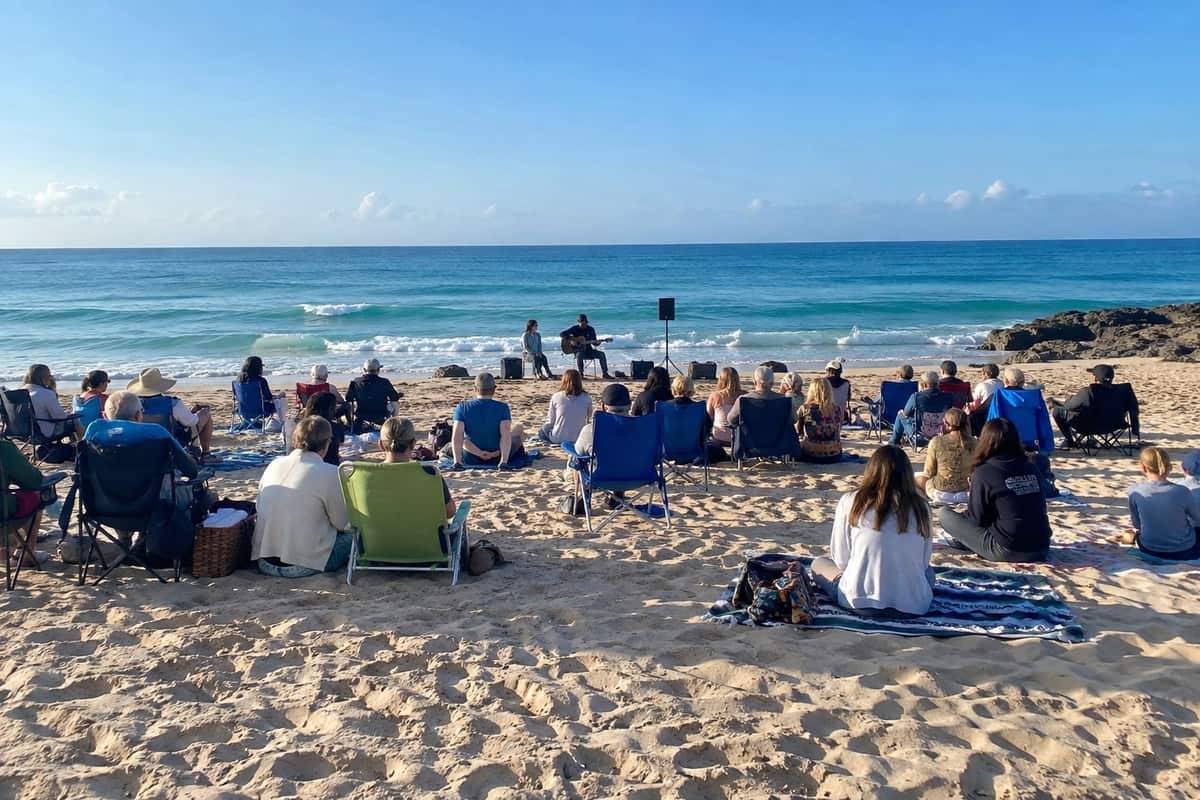 People on folding chairs and blankets on the sand, gathered for a Sunday morning service by the water